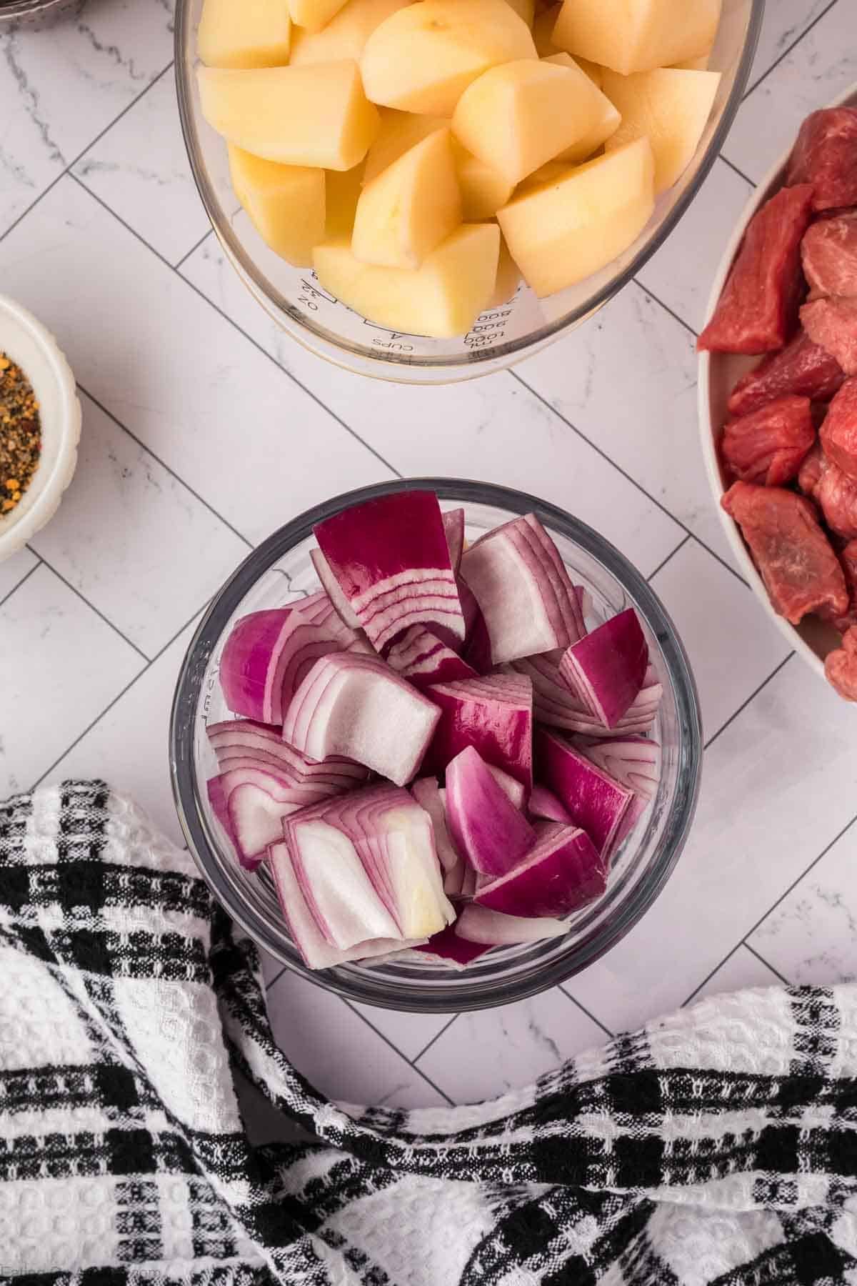 A bowl of chopped red onions sits on a checkered cloth, with a bowl of diced potatoes and raw beef nearby—perfect ingredients for a hearty crock pot steak and potato stew recipe, arranged on a white tiled surface.