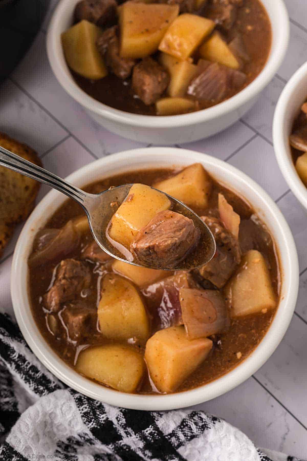 A close-up of a bowl of beef stew with chunks of beef, potatoes, and onions in broth. A spoon lifts a piece from the bowl. A black and white towel is beside this comforting crock pot steak and potato stew recipe.