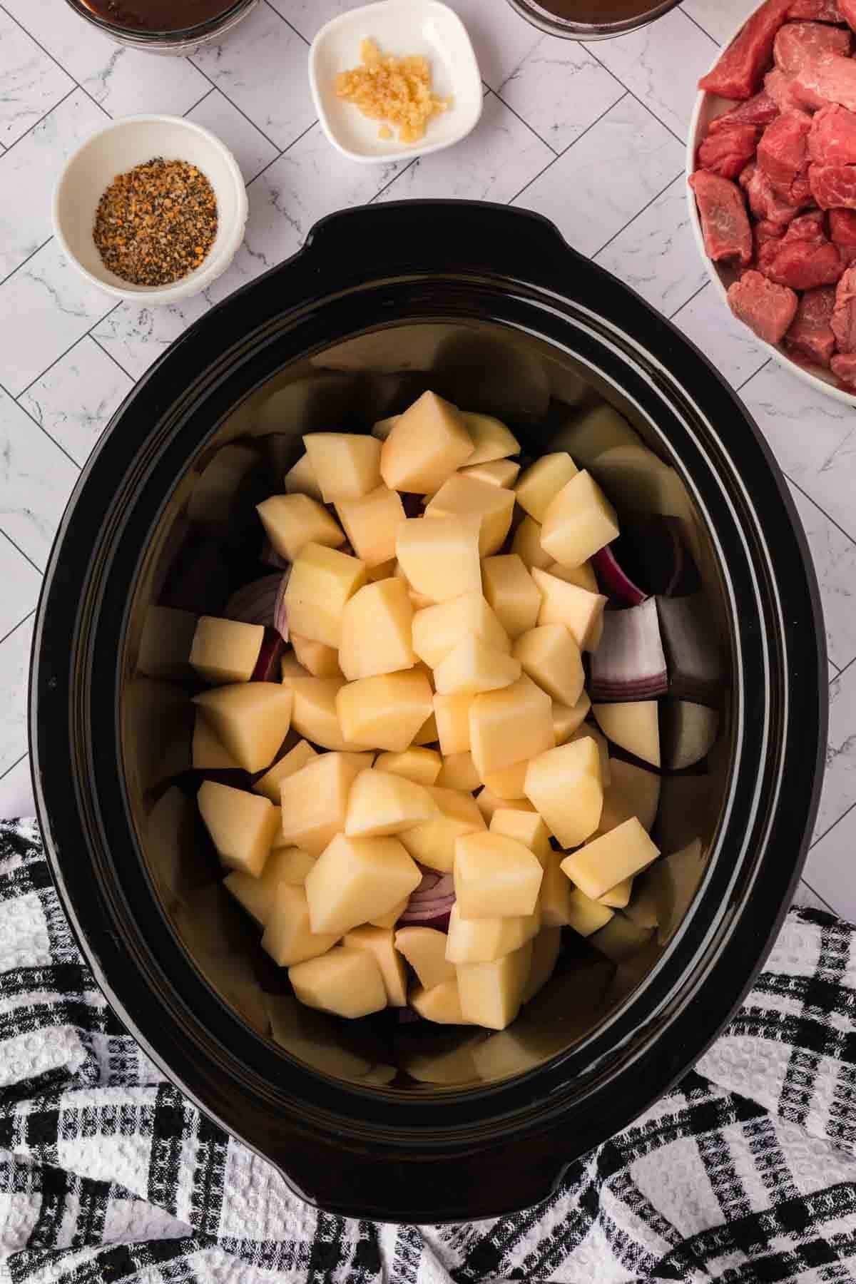 A crock pot steak and potato stew recipe comes to life as a slow cooker filled with chopped potatoes, onions, and cubed raw beef sits on the counter, surrounded by bowls of spices, minced garlic, and a checkered towel.