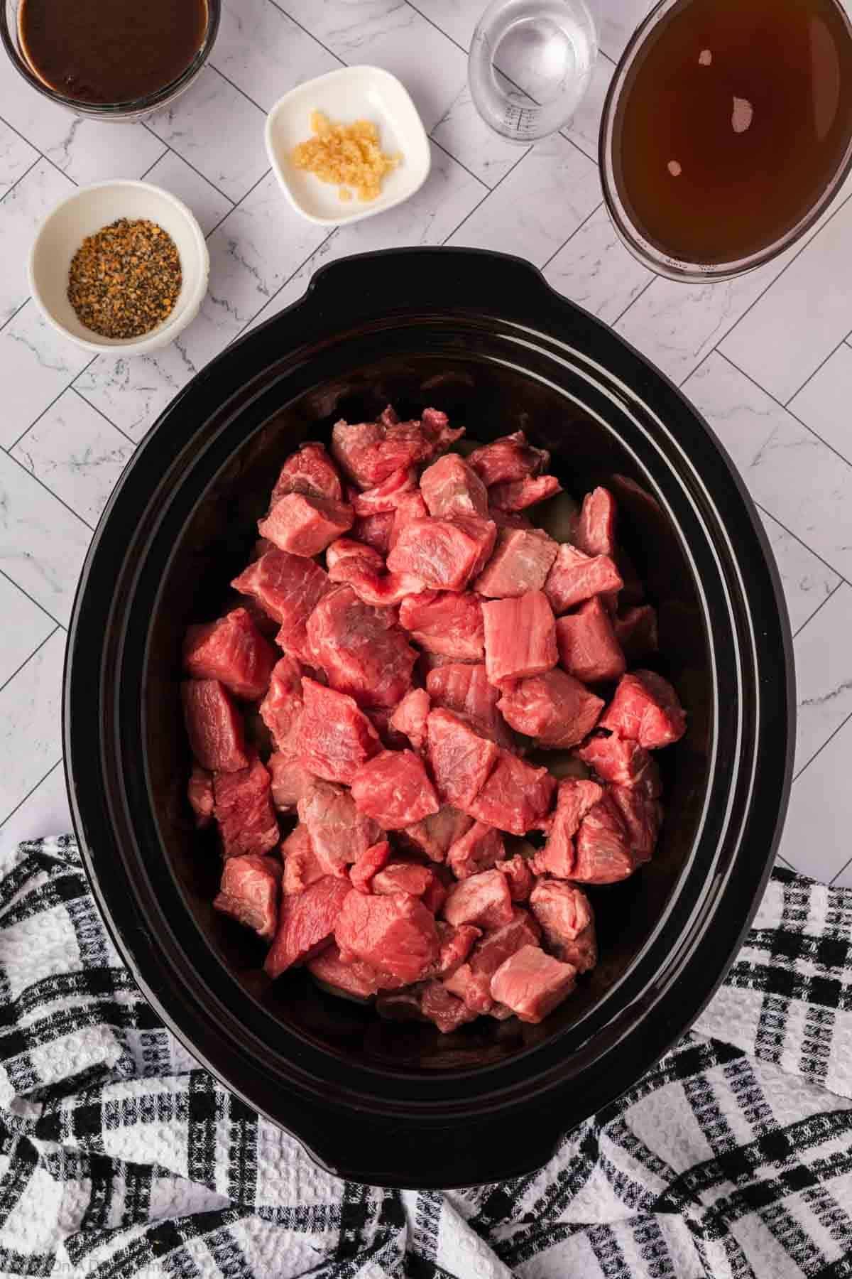 Top-down view of a crock pot steak and potato stew recipe in progress, with raw beef cubes in the slow cooker. Bowls of spices, minced garlic, broth, and dark sauce surround it on a white tiled surface with a black and white towel.