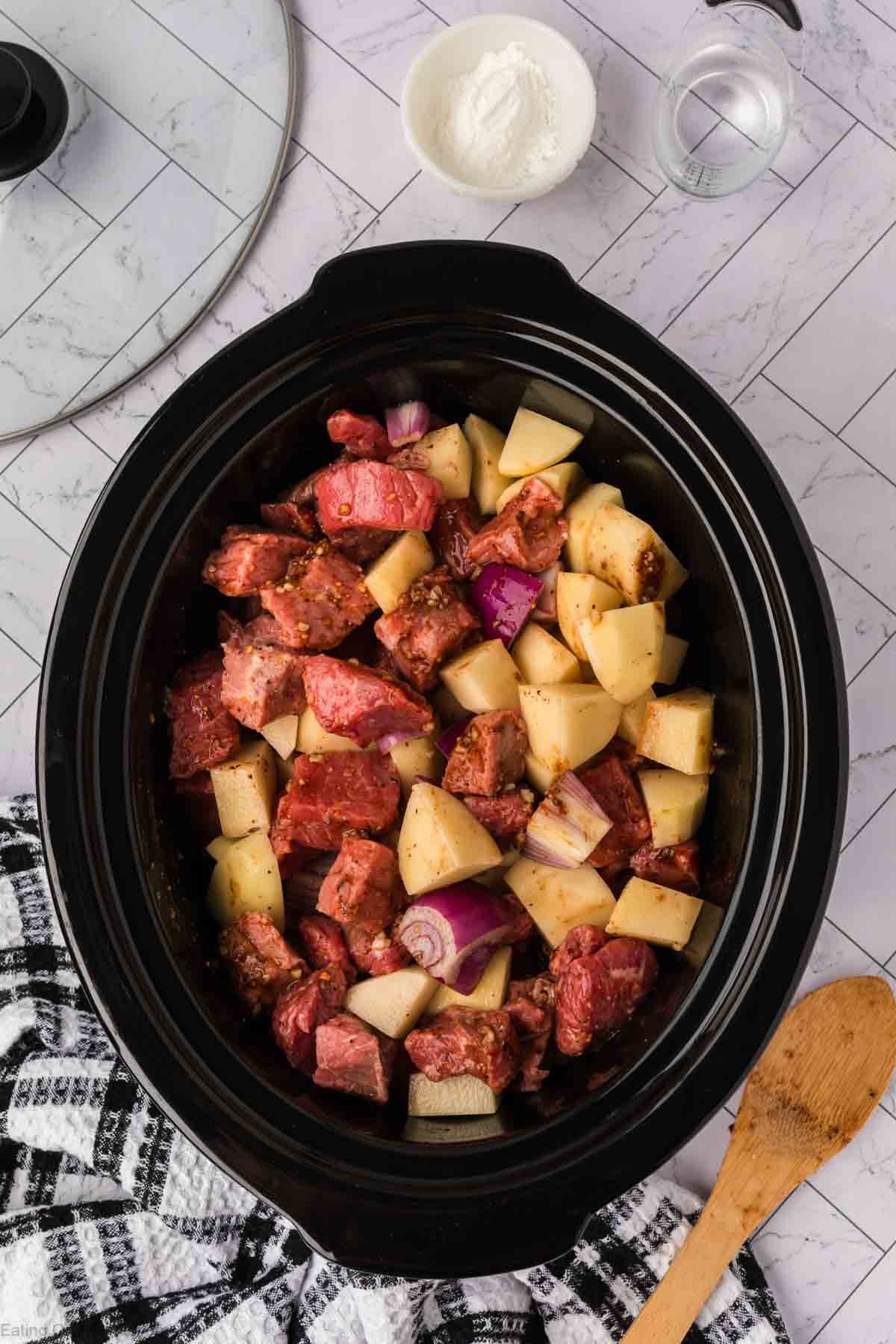 A crock pot steak and potato stew recipe takes shape in this image, with raw beef, potatoes, and red onions in the slow cooker, surrounded by a wooden spoon, black-and-white towel, flour bowl, and glass of water on a light tile surface.