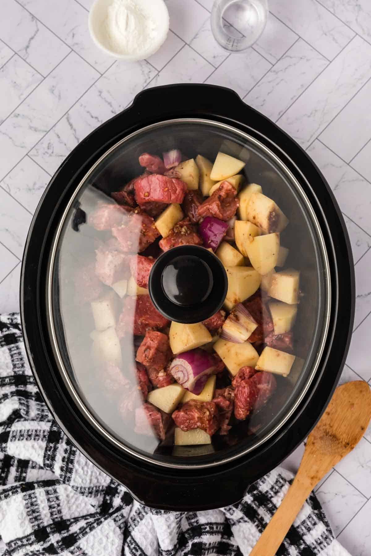 A crock pot steak and potato stew recipe features a slow cooker filled with chunks of beef, potatoes, and red onions on a checkered towel. A wooden spoon and bowls of ingredients rest nearby on the white tiled surface.