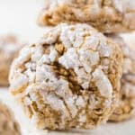 A close-up of a round pumpkin cake mix cookie coated in powdered sugar, its cracked surface revealing a soft, brown interior. Other similar cookies are blurred in the background.