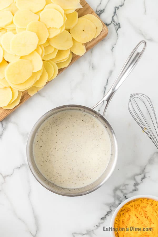 A stainless steel saucepan with creamy sauce sits on a marble countertop next to a small wire whisk. Behind the saucepan, a cutting board with thinly sliced potatoes and a bowl of shredded cheese hints at the delicious Cheesy Scalloped Potatoes Recipe being prepared. The text "Eating on a Dime .com" is in the bottom right corner.