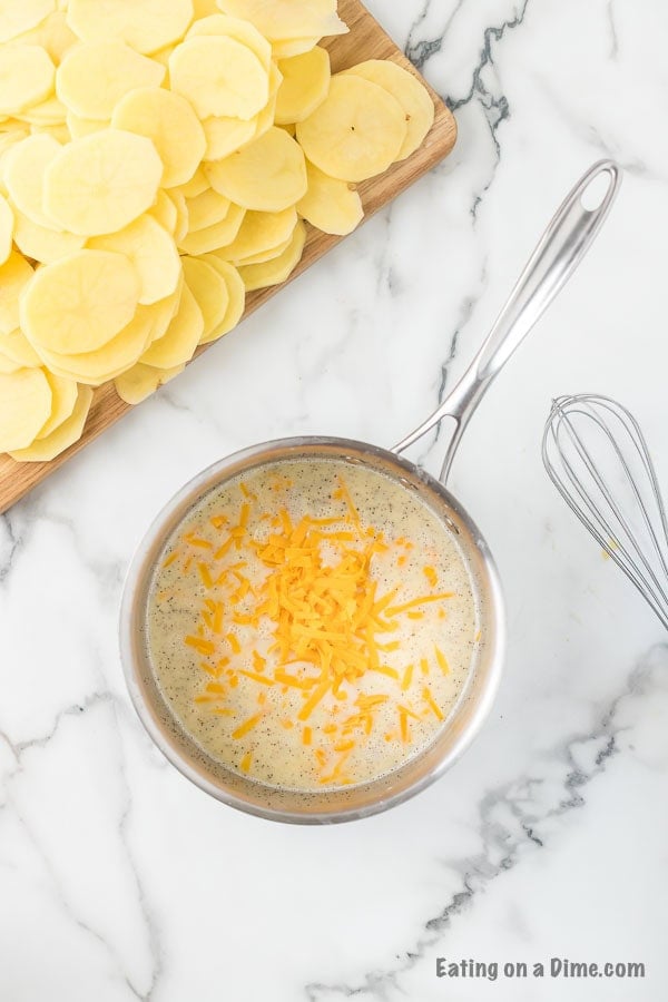 A saucepan filled with a creamy mixture topped with shredded cheese sits on a marble countertop, embodying the essence of a Cheesy Scalloped Potatoes Recipe. Beside the saucepan is a wooden cutting board with neatly sliced potatoes. A whisk rests nearby, and part of a website watermark is visible.
