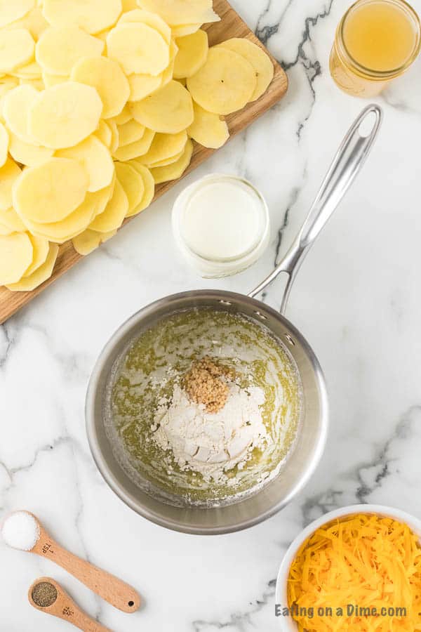 A marble countertop showcases ingredients for a Cheesy Scalloped Potatoes recipe. A saucepan contains flour and spices, with melted butter visible. Beside it, thinly sliced potatoes rest on a chopping board near a glass of milk, shredded cheese, a wooden spoon with pepper, and a dish of salt.