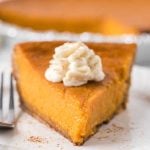 A close-up of a slice of Southern Sweet Potato Pie topped with a swirl of whipped cream, sitting on a white plate with a fork beside it. The rest of the pie is visible in the background.