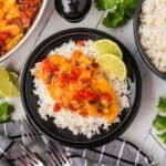 A black plate with white rice topped with cheesy Tex Mex Chicken Skillet Recipe and diced tomatoes, garnished with lime slices. A bowl of rice, cilantro, and lime wedges are nearby on a light surface with a black-and-white towel and forks.