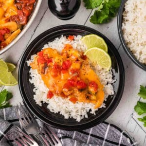 A black plate with white rice topped with cheesy Tex Mex Chicken Skillet Recipe and diced tomatoes, garnished with lime slices. A bowl of rice, cilantro, and lime wedges are nearby on a light surface with a black-and-white towel and forks.