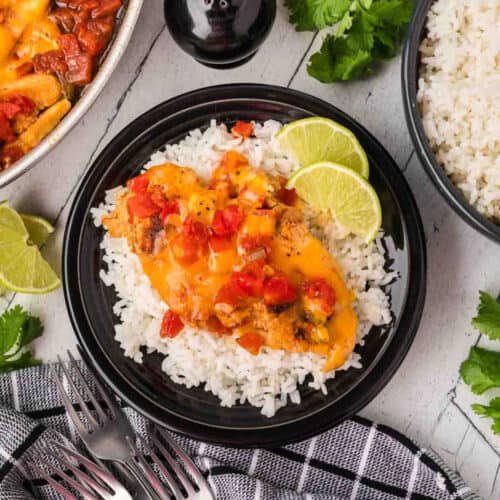 A black plate with white rice topped with cheesy Tex Mex Chicken Skillet Recipe and diced tomatoes, garnished with lime slices. A bowl of rice, cilantro, and lime wedges are nearby on a light surface with a black-and-white towel and forks.