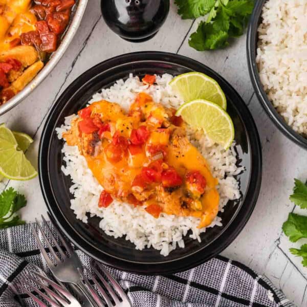 A black plate with white rice topped with cheesy Tex Mex Chicken Skillet Recipe and diced tomatoes, garnished with lime slices. A bowl of rice, cilantro, and lime wedges are nearby on a light surface with a black-and-white towel and forks.