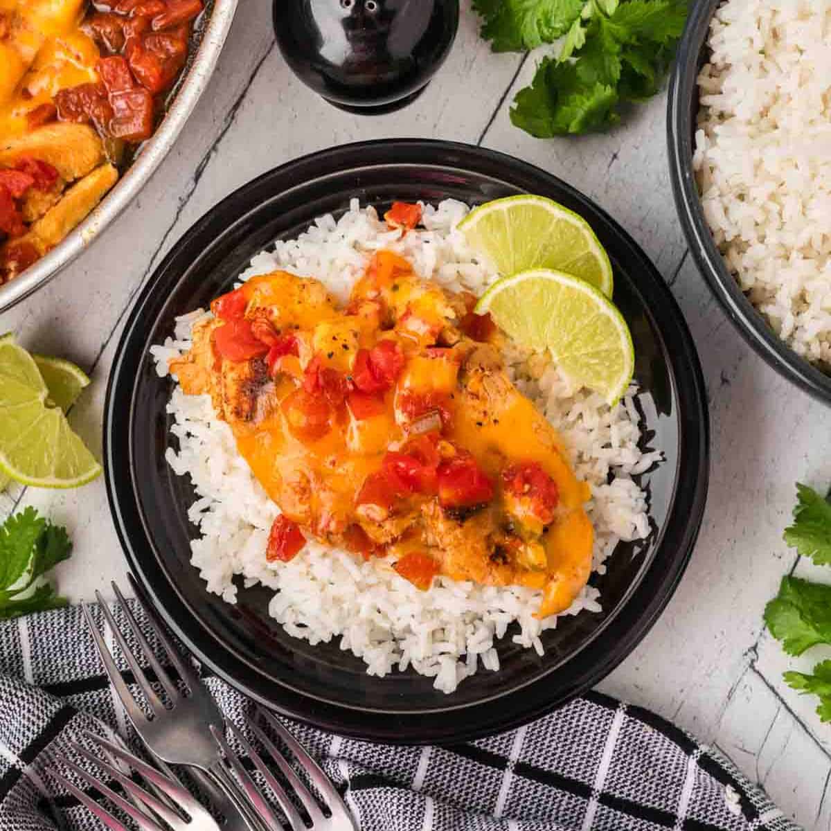 A black plate with white rice topped with cheesy Tex Mex Chicken Skillet Recipe and diced tomatoes, garnished with lime slices. A bowl of rice, cilantro, and lime wedges are nearby on a light surface with a black-and-white towel and forks.