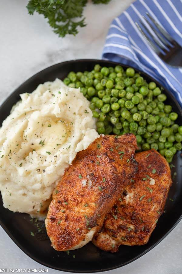 Close up image of pork chops on a black plate with a side of mashed potatoes and green peas. 