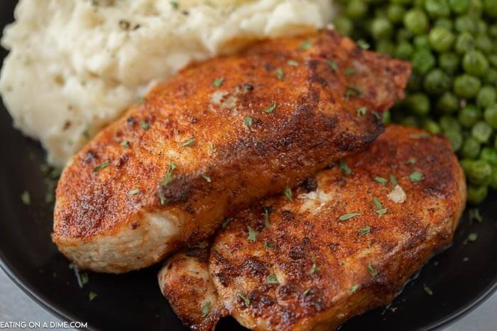 Close up image of pork chops on a black plate with a side of mashed potatoes and green peas. 