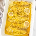 Four easy baked tilapia fillets in a white dish are topped with a yellow lemon-butter sauce, herbs, and pepper. Three lemon slices are placed on and around the fish. A checkered cloth is partially visible beside the dish.