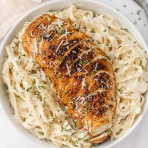 A bowl of creamy fettuccine Alfredo pasta topped with a seasoned, blackened chicken breast, garnished with shredded Parmesan cheese and chopped parsley.