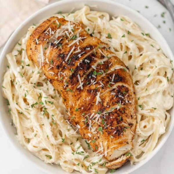 A bowl of creamy fettuccine Alfredo pasta topped with a seasoned, blackened chicken breast, garnished with shredded Parmesan cheese and chopped parsley.