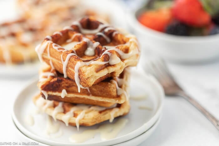 Cinnamon Roll Waffles stacked on top of each other with frosting drizzled on top with fruit and more waffles behind them.  