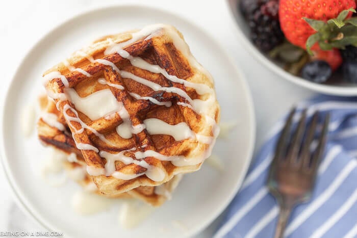Cinnamon Roll Waffles stacked on top of each other with frosting drizzled on top with a fork and fresh fruit next to it. 
