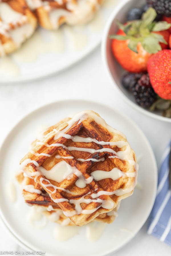 Cinnamon Roll Waffles stacked on top of each other with frosting drizzled on top with fresh fruit next to it. 