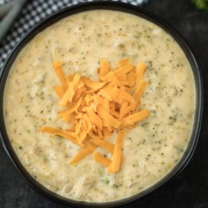 A bowl of creamy crockpot broccoli cheese soup topped with a generous pile of shredded cheddar cheese, set on a dark surface.