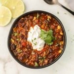 A hearty bowl of chili with black beans, corn, ground meat, and tomatoes—like a crockpot black bean soup with cilantro—topped with sour cream. Lime slices rest on a marble surface nearby.