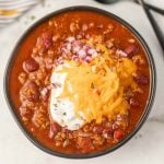 A bowl of Crockpot Cowboy Chili topped with shredded cheddar cheese, a dollop of sour cream, and chopped red onions, featuring ground meat, beans, and tomato pieces in a rich, red sauce.