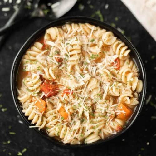 A black bowl filled with rotini pasta soup, visible pieces of carrots, tomatoes, shredded cheese, and sprinkled herbs on top—perfect for serving this Crock Pot Italian Chicken Soup recipe—set on a dark surface.