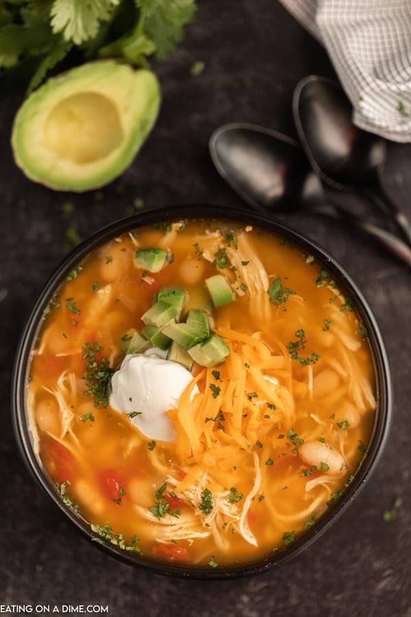 Close up image of a chicken taco soup in a black bowl with two black spoons and a avocado. 