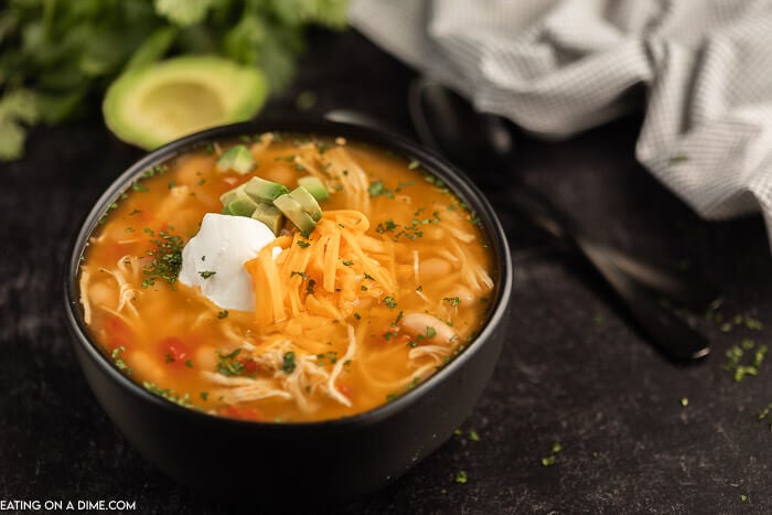 Close up image of chicken taco soup in a black bowl with two black spoons and a avocado. 