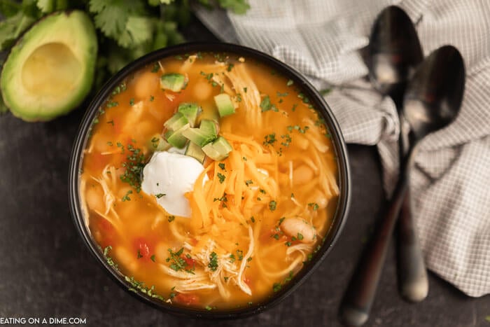 Close up image of chicken taco soup in a black bowl with two spoons and a avocado.