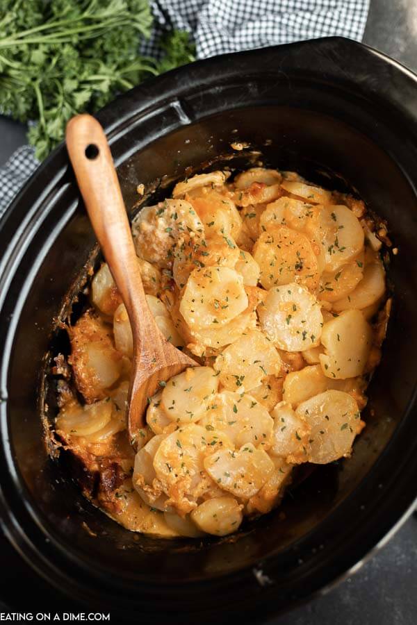 Close up image of scalloped potatoes in a crock pot with a wooden spoon. 