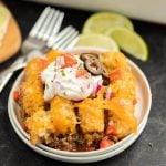 A plate of easy beef enchilada tator tot casserole recipe topped with melted cheese, sour cream, black olives, diced tomatoes, and red onions, served with lime wedges and two forks in the background.