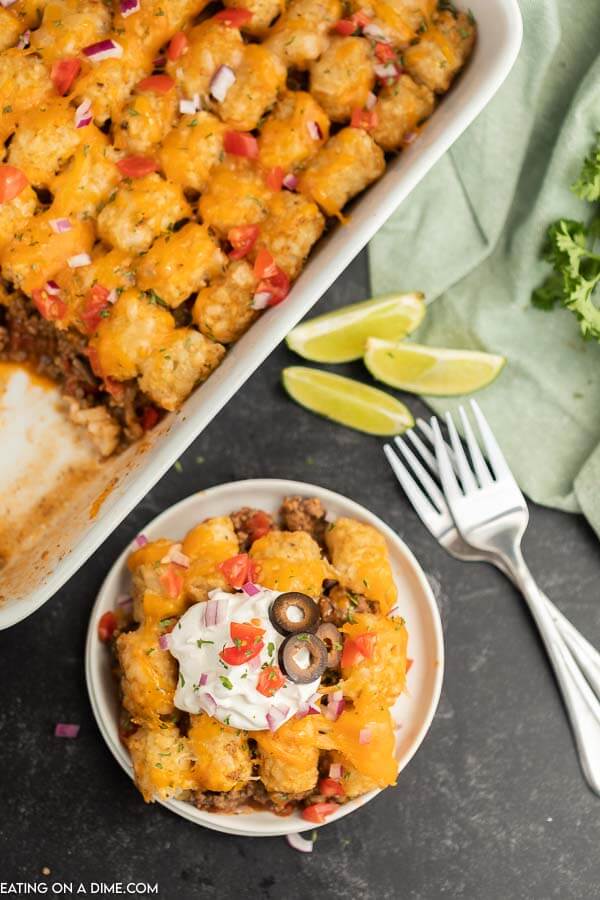 Close up image of beef enchilada tator tot casserole on a white plate. With a 9x13 pan of casserole