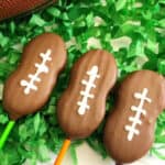 Three football cookie pops on sticks are coated in chocolate and decorated with white icing to resemble football laces, displayed on green shredded paper that looks like grass.
