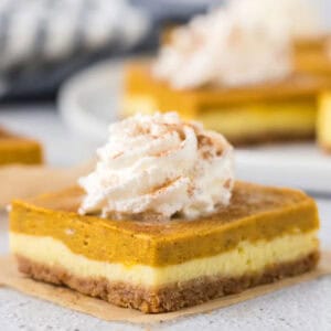 A close-up of Pumpkin Cheesecake Bars with three layers: a crumbly bottom crust, a creamy middle layer, and a spiced pumpkin topping, finished with a swirl of whipped cream and a dusting of cinnamon.