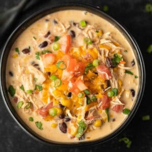 A bowl of creamy chicken soup, made with the Instant Pot Creamy Chicken Taco Soup Recipe, features black beans, corn, shredded chicken, diced tomatoes, cheddar cheese, and green onions, served on a dark surface.