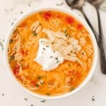 A bowl of creamy instant pot chicken taco soup topped with shredded cheese, a dollop of sour cream, crispy tortilla strips, and chopped herbs. Two spoons rest beside the bowl on a light surface.