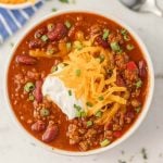 A bowl of Instant Pot Cowboy Chili topped with shredded cheddar cheese, a dollop of sour cream, and chopped green onions, featuring red beans and ground meat in a thick, red sauce.