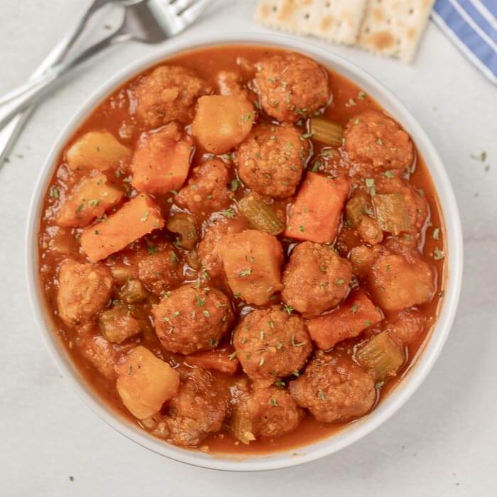 Close up image of a white bowl of meatball stew with a side of crackers. 
