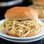 A shredded chicken sandwich piled high with tender chicken made from an Instant Pot Mississippi Chicken recipe, served on a round, soft bun atop a white plate.