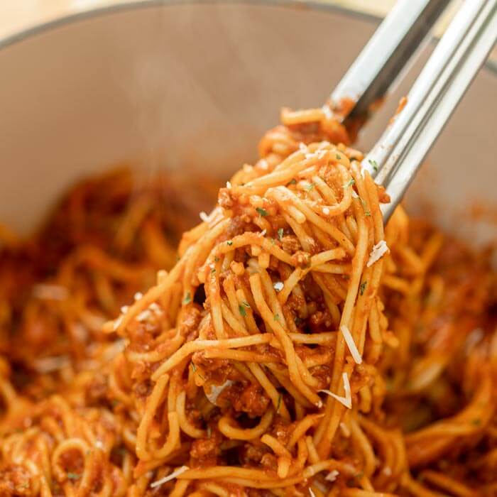 Close up image of a pot of spaghetti with a serving on silver tongs. 