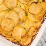 A close-up of a Cheesy Scalloped Potatoes Recipe in a white dish, showing golden, thinly sliced potatoes layered with creamy sauce and sprinkled with fresh herbs. The edges are perfectly browned and crispy.