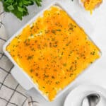 A square white baking dish filled with Cheesy Mashed Potatoes, topped with melted cheddar cheese and chopped chives, sits on a light surface next to a checkered towel, parsley, and a spoon.