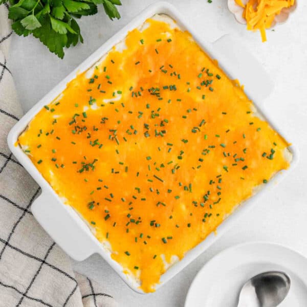 A square white baking dish filled with Cheesy Mashed Potatoes, topped with melted cheddar cheese and chopped chives, sits on a light surface next to a checkered towel, parsley, and a spoon.