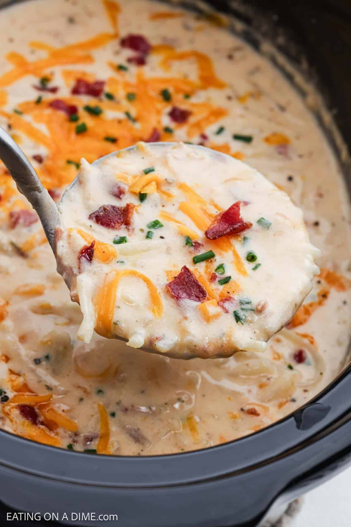 Ladle of creamy soup of a crock pot of soup topped with shredded cheese and crispy bacon.