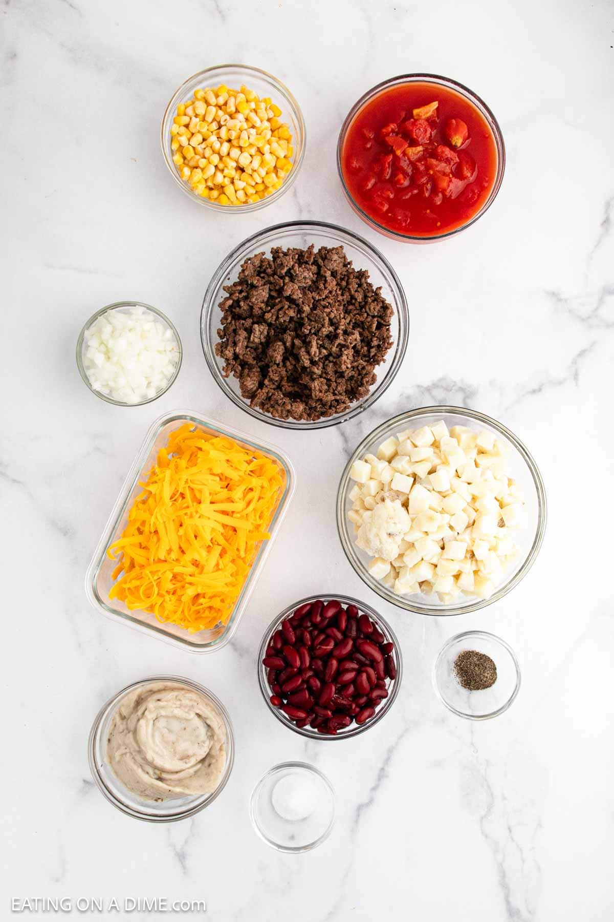 Nine bowls on a white surface display the ingredients for a classic Crockpot Cowboy Casserole: corn, diced tomatoes, ground beef, chopped onions, shredded cheddar cheese, diced potatoes, kidney beans, cream soup, salt, and pepper.