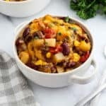 A white bowl filled with Crockpot Cowboy Casserole featuring ground beef, kidney beans, corn, diced potatoes, and tomatoes, topped with parsley. A checked napkin and fresh herbs are nearby.