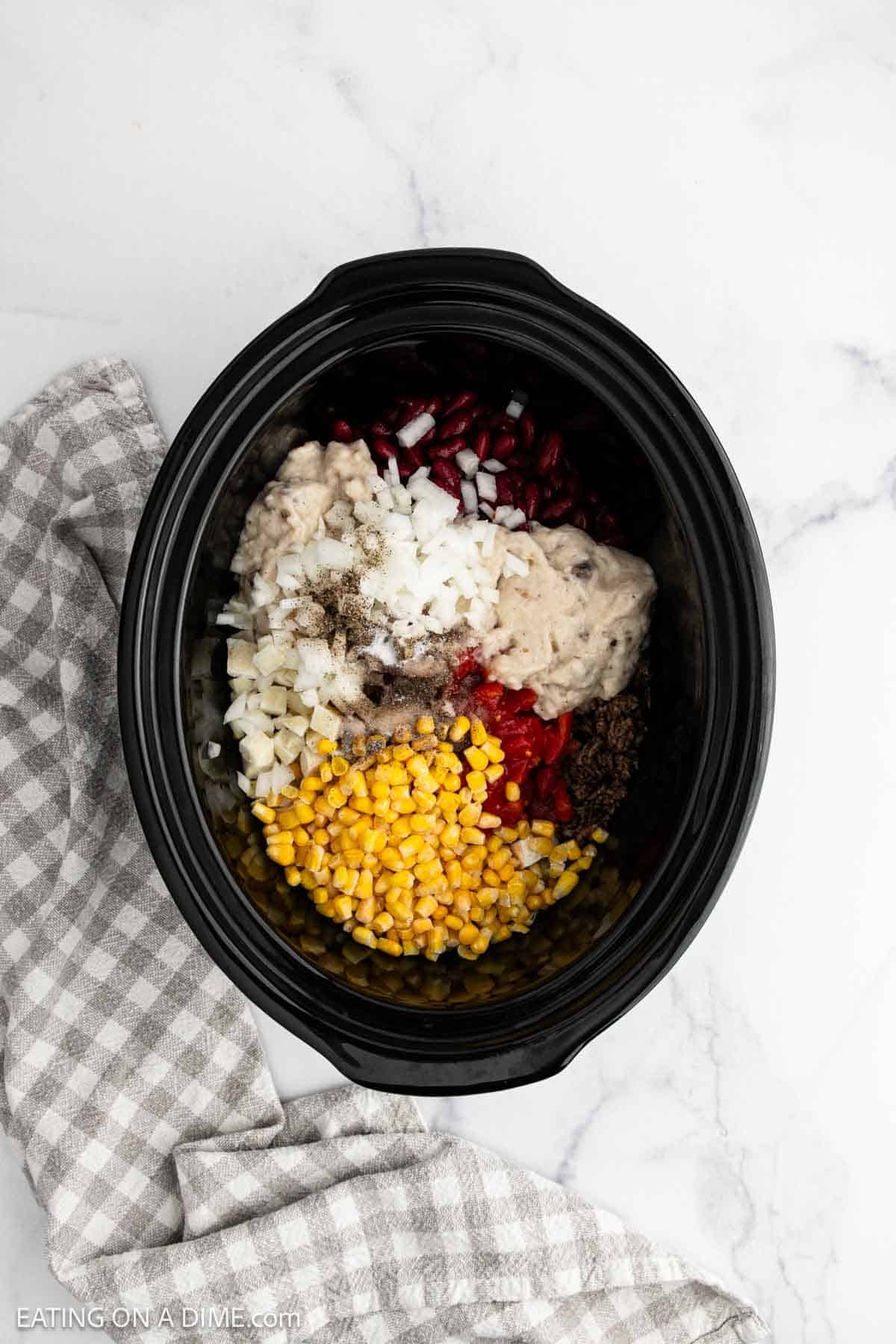 A Crockpot Cowboy Casserole filled with ground beef, corn, diced onions, red beans, tomatoes, and a creamy soup base sits on a white marble surface next to a gray checkered kitchen towel.