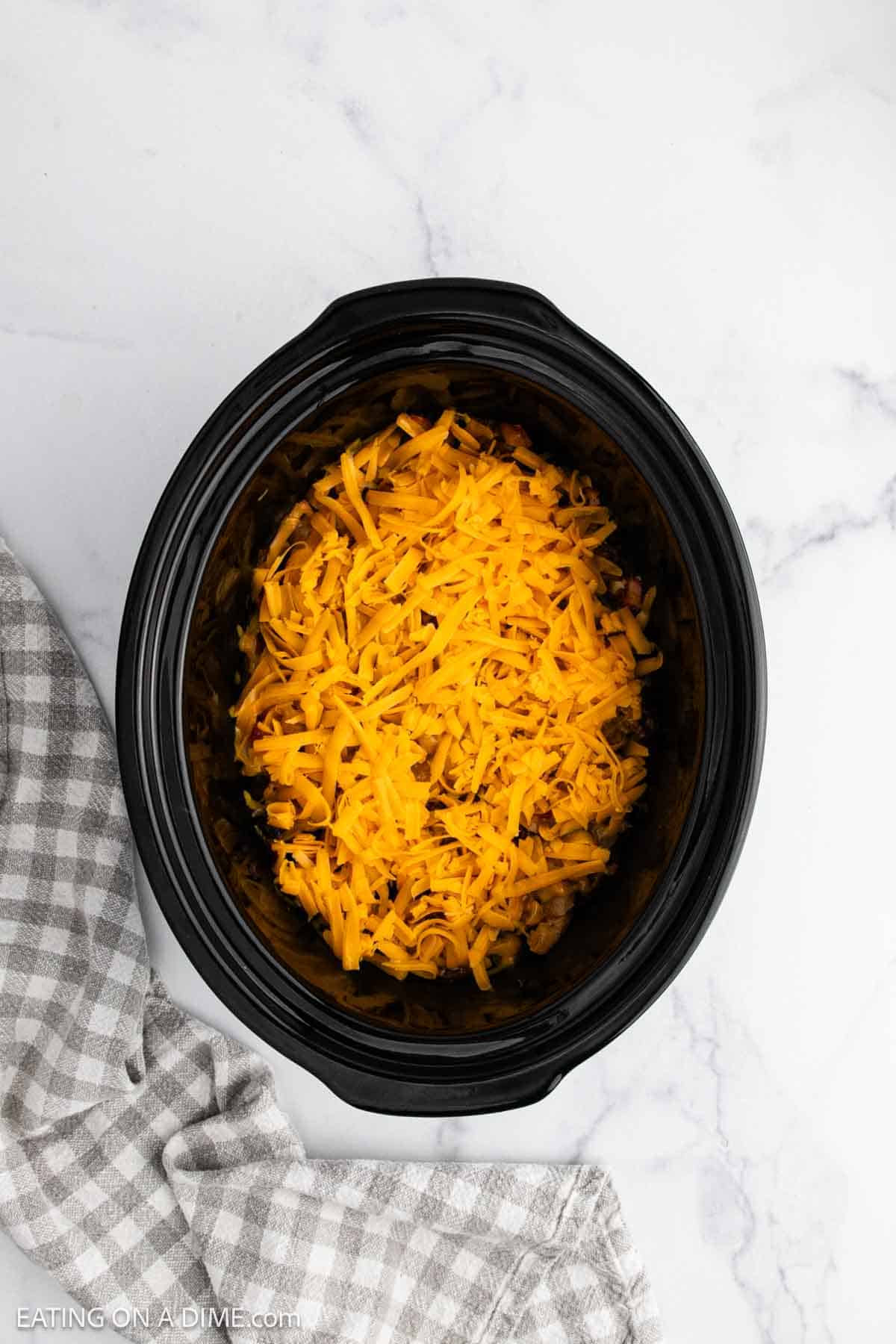 A black slow cooker filled with shredded cheddar cheese sits on a white marble surface next to a gray and white checkered towel, ready for making a hearty Crockpot Cowboy Casserole.